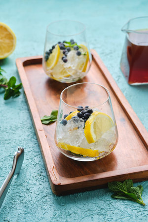 Preparing refreshing drink, iced tea with lemon slices, mint and blueberries in glasses on turquoise concrete background. Soft drinks. Summer drinksの写真素材