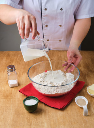 Step by step preparation of yeast dough with milk. Woman pouring milk into a bowl with flour on a wooden background. Cook's work in the kitchen, baking. Step by step recipesの写真素材