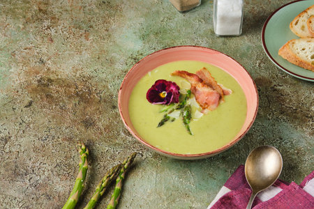 Fresh asparagus puree soup with asparagus pieces and fried bacon in a pink ceramic plate on a green concrete background. Recipes asparagus. Seasonal vegetables.の写真素材