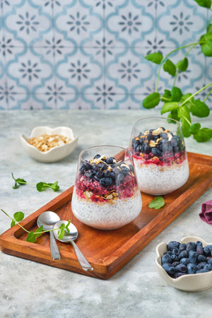 Chia pudding with nut granola, honeysuckle berry puree and whole honeysuckle berries in transparent glasses on gray concrete background. No bake desserts, desserts in glasses. Healthy foodの写真素材