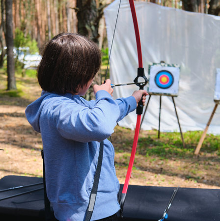 A teenager in a blue hoodie shoots a target with a sport bow at a shooting range in the forest. Active recreation, sports. Sport shootingの写真素材