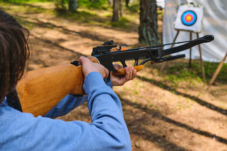 Teenager in blue hoodie shoots from black sport crossbow at target at shooting range in forest. Active recreation, sport. Sport shooting with crossbowの写真素材