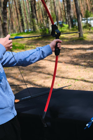 A teenager in a blue hoodie shoots a target with a sport bow at a shooting range in the forest. Active recreation, sports. Sport shootingの写真素材