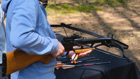 Teenager in blue hoodie loading black sport crossbow at shooting range in forest. Active recreation, sport. Sport shooting with crossbowの写真素材