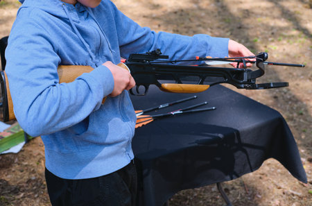 Teenager in blue hoodie loading black sport crossbow at shooting range in forest. Active recreation, sport. Sport shooting with crossbowの写真素材