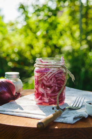 Pickled sliced ââred onion in a clear glass jar with marinade on a table in the yard. Grill season. Appetizer or seasoning for meat or fish dishesの写真素材