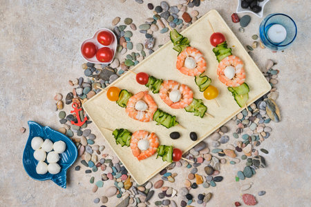 Skewers with shrimps, cherry tomatoes, mozzarella cheese and cucumber slices on a rectangular plate served in a marine style on a light concrete background. Seafood recipes.の写真素材