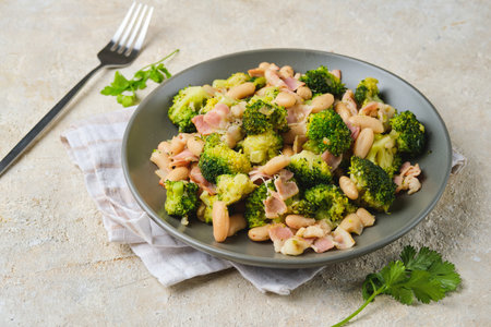 Fried broccoli with bacon and canned white beans on a gray plate against a light concrete background. Broccoli recipes. Vegetable dishes.の写真素材