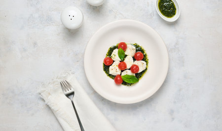 Winter caprese salad with marinated cherry tomatoes, mozzarella, and pesto sauce on a light plate against a white concrete background. Salad recipes.の写真素材