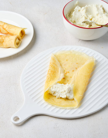 Step-by-step instructions for making dessert pancakes with cottage cheese filling on a light concrete background. Step three: wrapping the cottage cheese filling into the pancake. Step-by-step recipesの写真素材