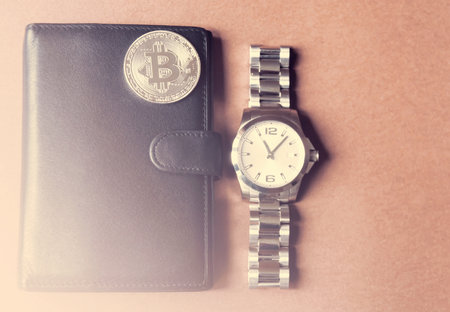 On a brown background is a black wallet, on it is a silver coin bitcoin, beside a man's watch. With a lighting.の写真素材