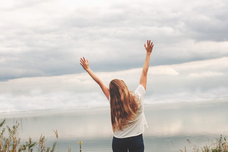 Beautiful girl on the lake. The view from the back. Hands up. Cloudy sky, sunset.