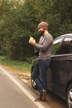 Young guy eating a burger near a car on an empty road. Food on the trip. Food on the go. Autumn...