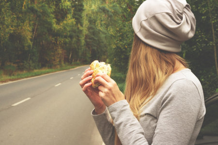 A young woman is eating a burger near a car on an empty road. Food on the trip. Food on the go. Autumn travel. Fast foodの写真素材