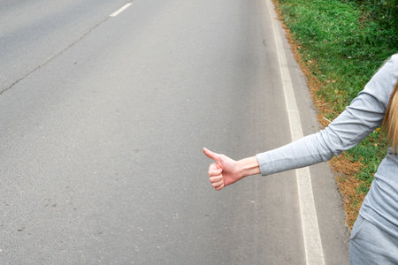 A young and beautiful girl votes on the road. Autumn road. Catch a car on an empty road. Hitchhiking. Free travel by carの写真素材