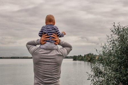 A child on his fathers neck. Walk near the water. Baby and dad against the sky