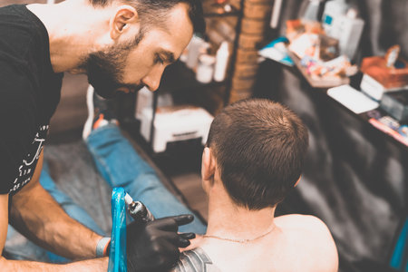 Tattoo salon. The tattoo master is tattooing a man on his shoulder. Tattoo machine, safety and hygiene at work. Close-up, tinted, tattooistの写真素材