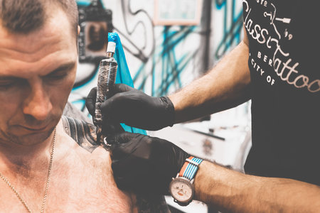 Tattoo salon. The tattoo master is tattooing a man on his shoulder. Tattoo machine, safety and hygiene at work. Close-up, tinted, tattooist.の写真素材