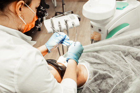 Dentist young woman treats a patient a man. The doctor uses disposable gloves, a mask and a hat. The dentist works in the patients mouth, uses a professional tool.の写真素材