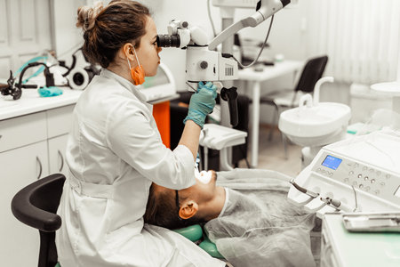 Dentist young woman treats a patient a man. The doctor uses disposable gloves, a mask and a hat. The dentist works in the patients mouth, uses a professional tool.の写真素材