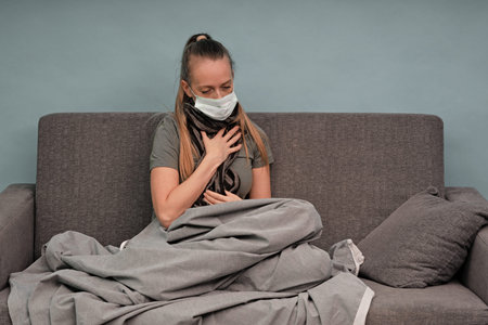 The girl is sick at home. Sitting on a couch, wrapped in a blanket, a scarf around his neck. Sore throat and chest. Uses a face shield. Coronavirus.の写真素材