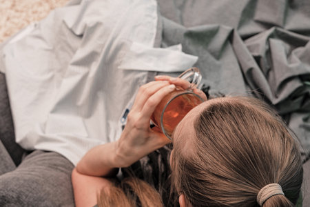 The girl is sick at home. Sitting on a couch, wrapped in a blanket, a scarf around his neck. Drinks hot tea. View from above. Coronavirus.の写真素材