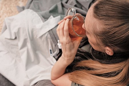 The girl is sick at home. Sitting on a couch, wrapped in a blanket, a scarf around his neck. Drinks hot tea. View from above. Coronavirus.の写真素材