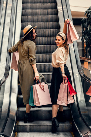 Two girls on an escalator in a shopping center. In the hands of paper bags. Shopping, leisure and entertainment.の写真素材