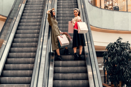 Two girls on an escalator in a shopping center. In the hands of paper bags. Shopping, leisure and entertainment.の写真素材