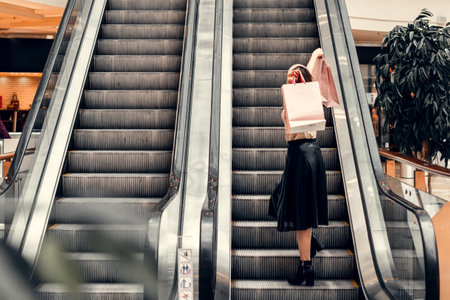 Two girls on an escalator in a shopping center. In the hands of paper bags. Shopping, leisure and entertainment.の写真素材