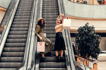 Two girls on an escalator in a shopping center. In the hands of paper bags. Shopping, leisure and entertainment.の写真素材