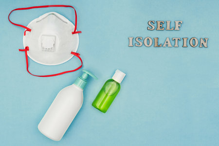 Coronavirus, Covid-19. Inscription self-isolation, a protective mask, an antiseptic and a dispenser for living soap on a blue background. Self-isolation and quarantine, personal protective equipment.の写真素材