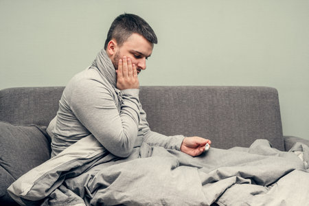 Disease. Cheer at home. A young man is sick, is treated at home. Blows her nose into a napkin, runny nose. Body temperature measurement. Infection, epidemic, bacillus carrier. Coronavirusの写真素材