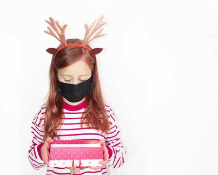 quarantined Santa deer, Beautiful girl red sweater Christmas, and New year gift. The happy small girl in Santa s hat has a Christmas. in a protective black mask on a white background banner, Christmas and covid-19 concept. High quality photoの写真素材