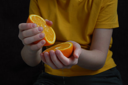 A boy in a yellow t-shirt holds orange slices in his hands, squeezes orange juice from one slice on a dark backgroundの写真素材