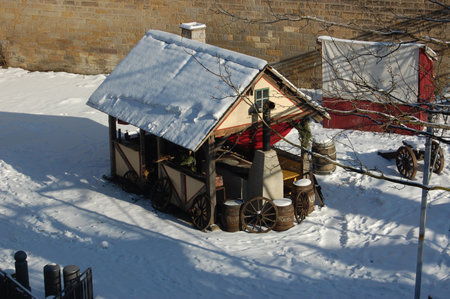 a wooden house with a chimney on a background of snow, barrels and wheels from an old cart are nearby against the background of an old brick wallの写真素材