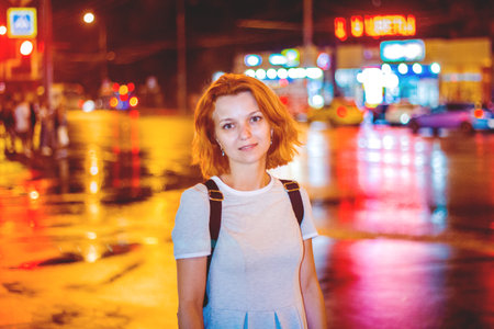 cute fiery red-haired girl standing in the night city lit with lights and headlights passing cars.の写真素材