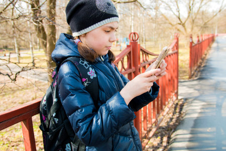 schoolgirl with backpack in warm sunny weather stands at the fence and looks at the phone.