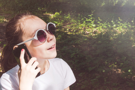 Happy little girl with sunglasses looking at the sun in the summer forest and talking on mobile device.の写真素材