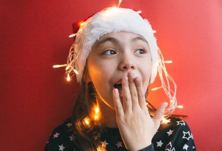 Cute little girl in Santa Claus hat with garland on red background.の写真素材
