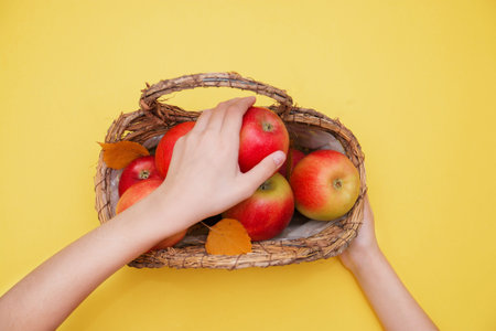 child hands holding basket with red apples on yellow background autumn composition and concept.