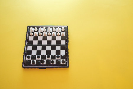 small pocket chess, plastic chess pieces placed on a chessboard on a yellow background top view.