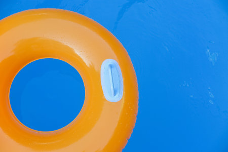 Children inflatable orange swimming ring with white handles floating in blue pool. Top view,...