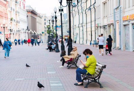 Unknown people in a mouth mask walk in a public place, sit on benches during the quarantine. Moscow, Russia, May 2020.のeditorial素材