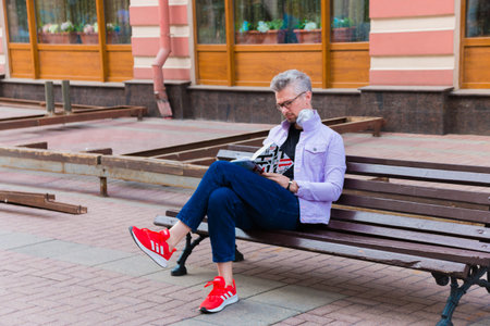 Unknown man in a mouth mask against smog air pollution with PM2.5 sitting on a bench and reading a book, walking on a schedule. Moscow, Russia, May 2020.のeditorial素材