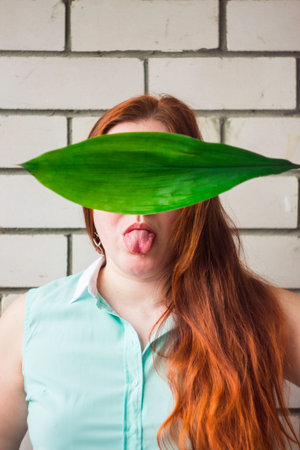 girl with red long hair and protruding tongue covers her face with a green leaf against a brick wall...