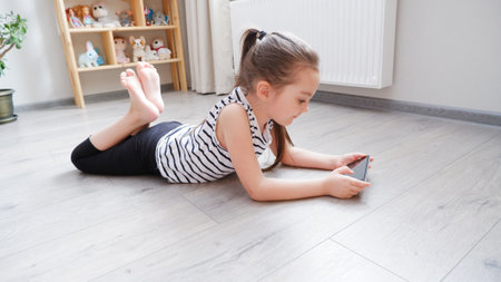 Distance learning. Little girl lying on a wooden floor with a phone, watching a cartoon or making a video call on a computerの写真素材