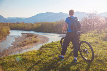 Boy cyclist standing on a mountain and looks at the river at sunset Concept for travelers. View from...