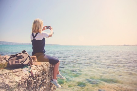 young girl traveler takes pictures on the camera beautiful view from the sea. The girl loves to travel Concept for travelers. View from back of the tourist traveler on background seaの写真素材