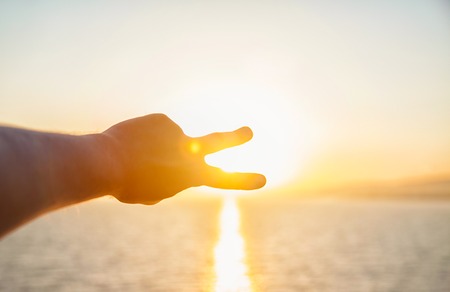 The man's hands stretch toward the sun on the sea. A man looks at the sunset through the folded fingers.の写真素材